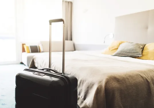 Koffer mit ausgezogenem Griff steht neben einem gemachten Bett in hellem Hotelzimmer.
