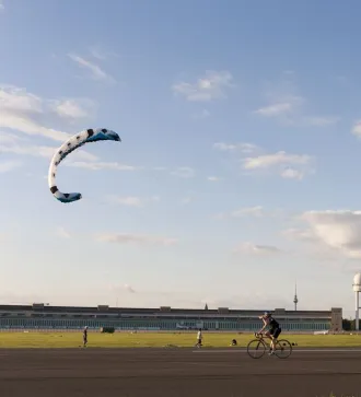 Person fährt Fahrrad auf weitem Feld, bunter Drachen fliegt am blauen Himmel.