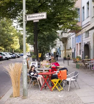 Menschen sitzen an bunten Tischen vor einem Straßencafé an einem sonnigen Tag.