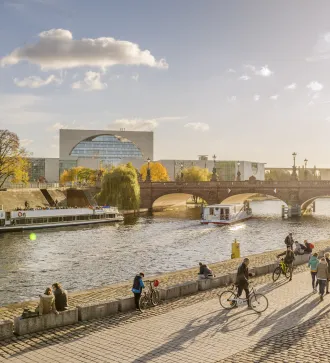 Menschen spazieren am Spreeufer, vorbeifahrende Boote, Brücke und modernes Gebäude im Sonnenlicht.