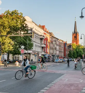 Belebte Straßenszene mit Radfahrern, Fußgängern und Kirche im Hintergrund bei Tageslicht.