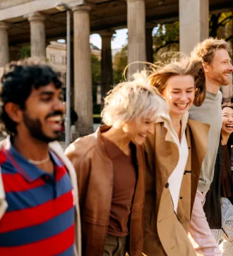 Fünf lachende Menschen spazieren gemeinsam an sonnigem Tag vor Säulengang.