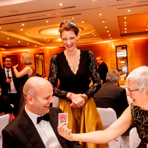Elegant gekleidete Menschen sitzen lachend an einem festlich gedeckten Tisch im Ballsaal.