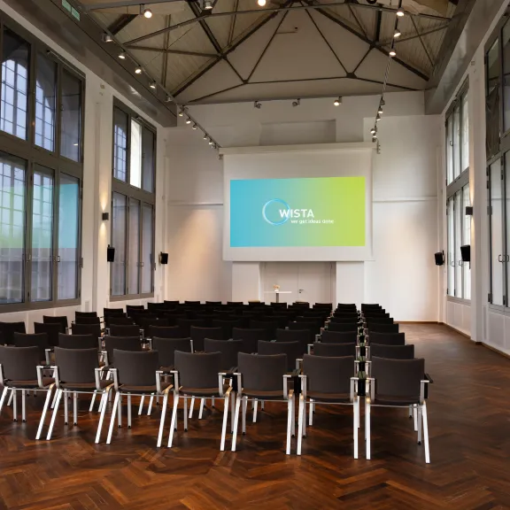 Leerer Konferenzraum mit Stuhlreihen und großer Leinwand vor hohen Fenstern