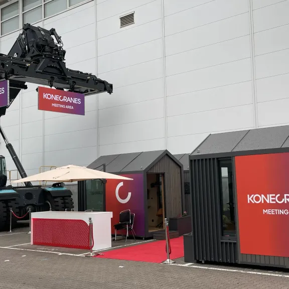 Außenbereich mit Containern, Messestand und großem Gabelstapler vor einer Halle.