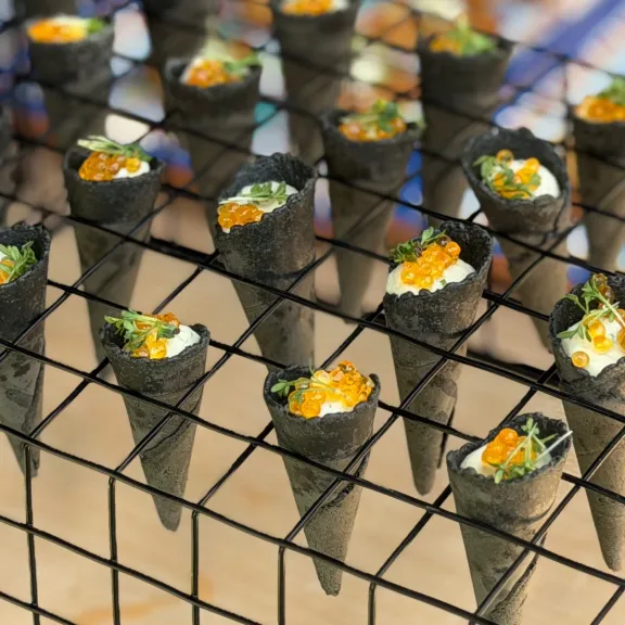 Mehrere kleine Sushi-Handrollen mit Lachsrogen auf einem Metallgitter angerichtet.