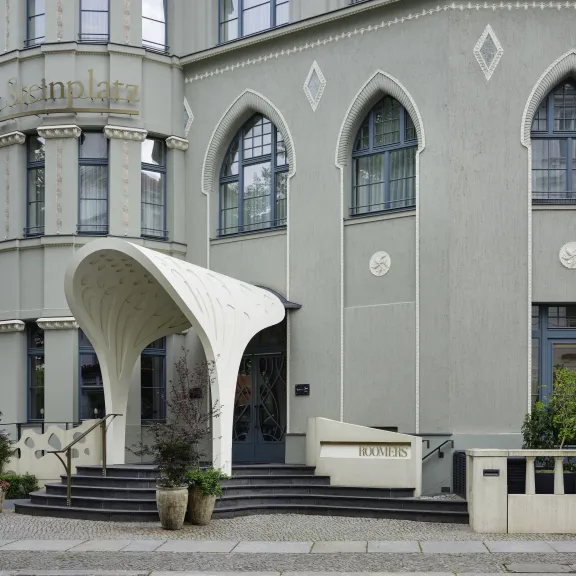 Gebäude mit rundem Vordach, großen Fenstern und Blumenkübeln am Eingang.
