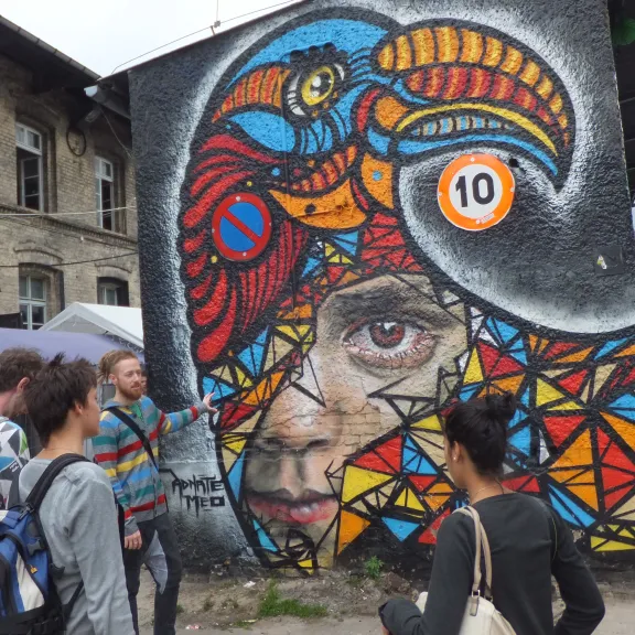 Bunte Graffiti-Wand mit Vogel- und Frauengesicht, mehrere Menschen davor.