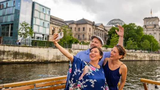Drei Menschen machen ein Selfie auf einem Boot vor Berliner Gebäuden am Fluss.