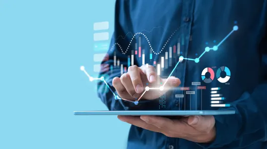 Person using a tablet with digital financial graphs and charts projected above it.