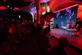 Publikum sitzt in dunklem Saal vor rot beleuchteter Bühne mit zwei Personen.