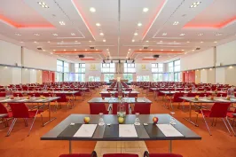 Großer, heller Konferenzsaal mit roten Stühlen und gedeckten Tischen.
