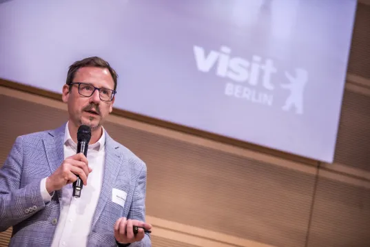 Man holding a microphone speaking, "visit Berlin" projected on screen behind him.