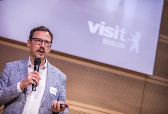 Mann mit Mikrofon präsentiert vor „visit Berlin“-Logo.