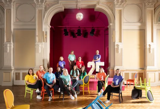 Gruppe von Menschen sitzt vor einer Bühne mit der Zahl 25, bunte Stühle, festlicher Raum.