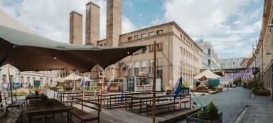 Outdoor seating area with wooden tables, tents, and tall industrial buildings in the background.