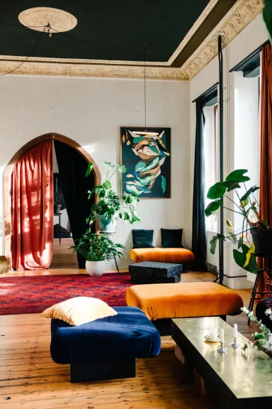 Cozy living room with colorful furniture, plants, and sunlight streaming through large windows.
