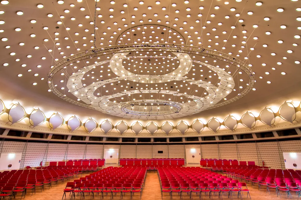 Großer Saal mit roten Stuhlreihen und beeindruckender, beleuchteter Decke.